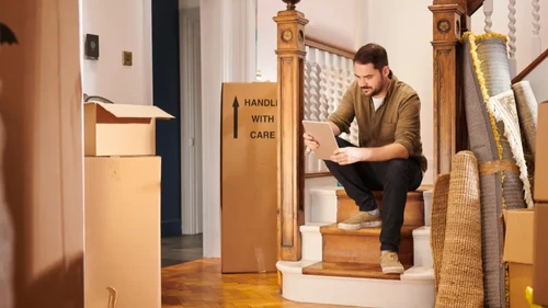 Man sitting on stairs looking at a clipboard and surrounded by moving boxes